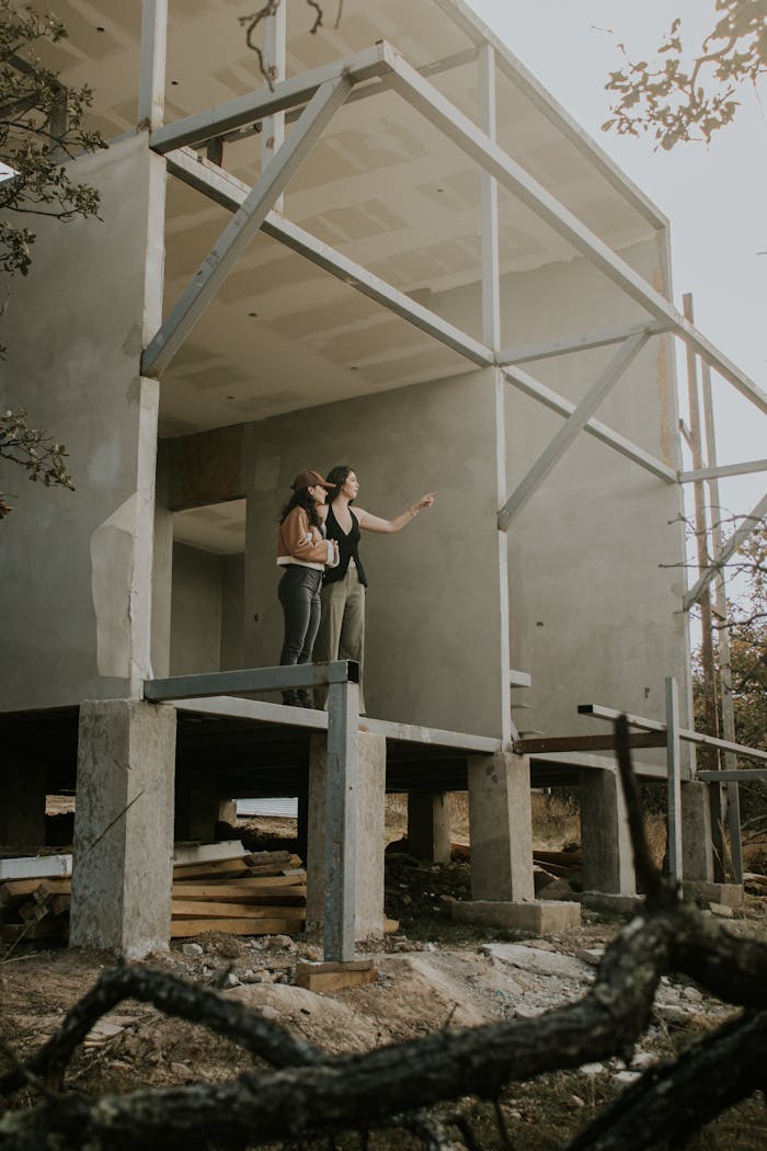 Two adults observe a modern concrete house under construction, surrounded by nature.