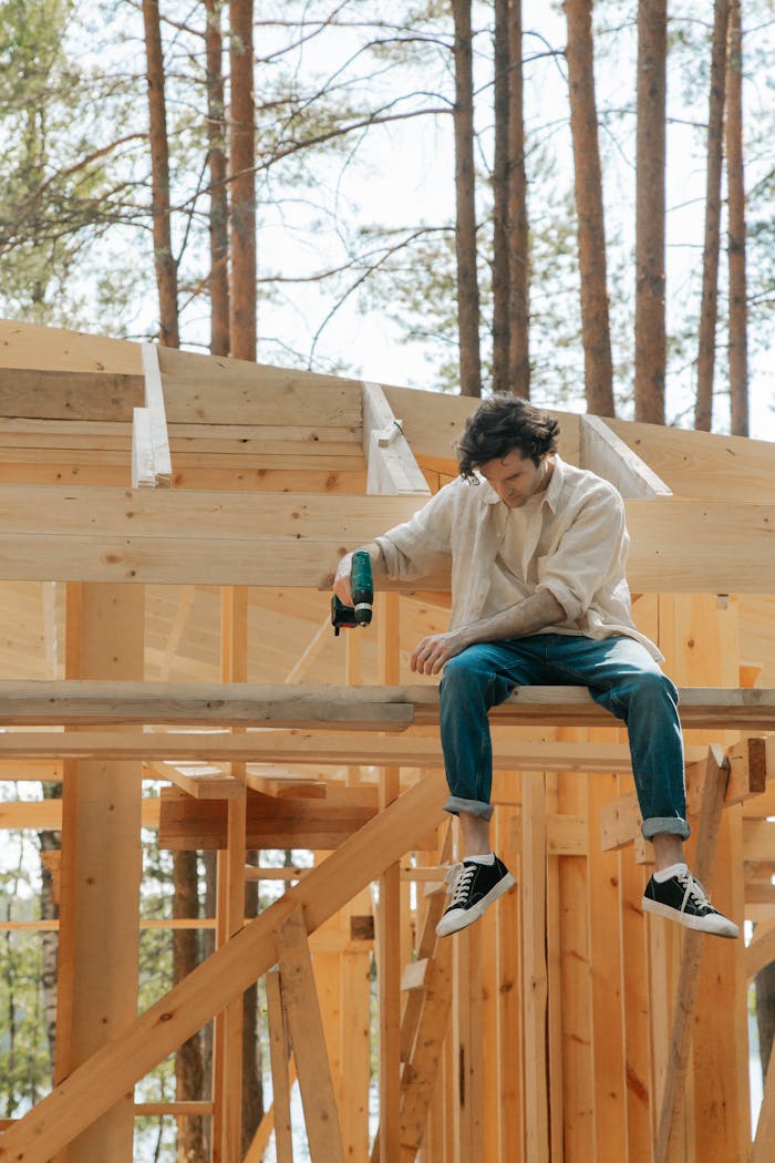 A young man using a cordless drill on a wooden house under construction in a forest setting.