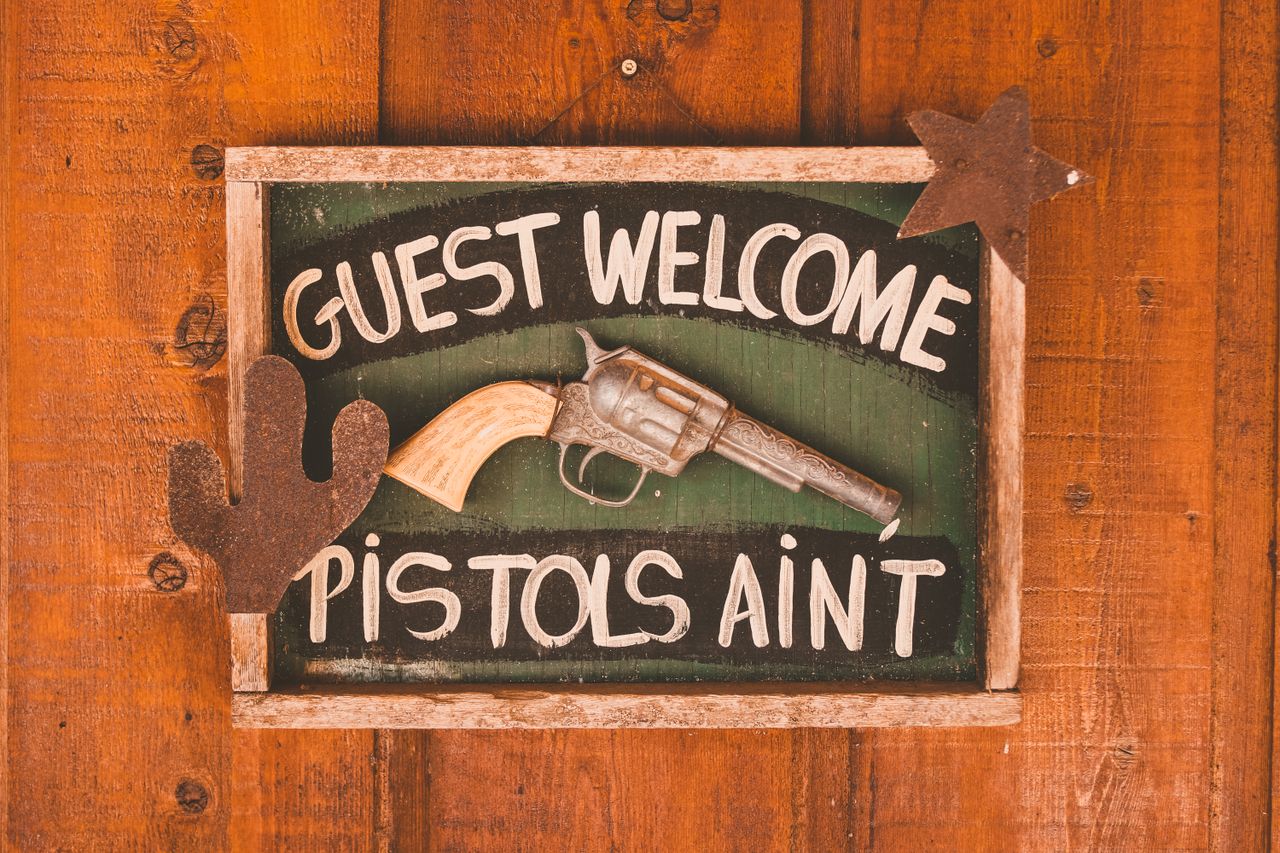 our-story Framed rectangular signboard with inscription Guest Welcome Pistols Aint decorated with artificial gun and silhouettes of cactus and star on wooden door